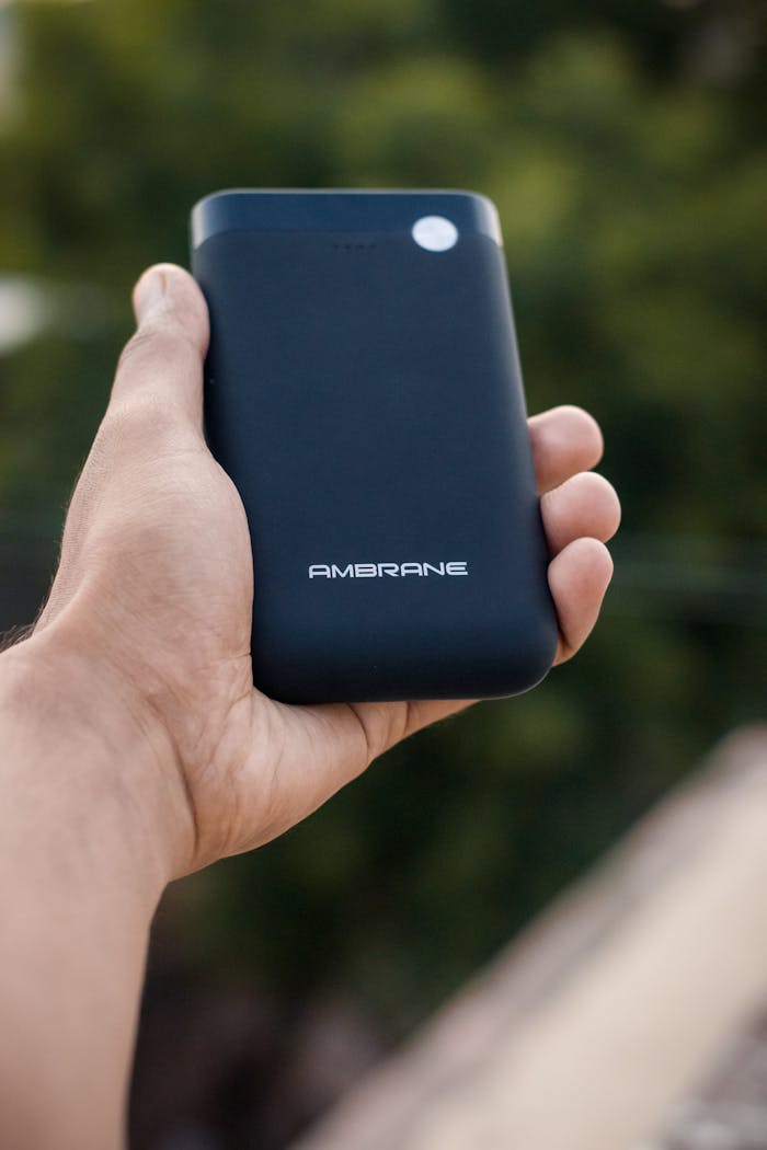 services-06 Close-up of a hand grasping a sleek black power bank with AMBRANE branding, set against an outdoor background.